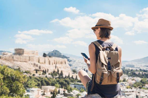 A person wearing a hat and backpack sits on a rock, using a smartphone while planning their day trip. In the background, the Acropolis of Athens stands majestically under a clear blue sky.